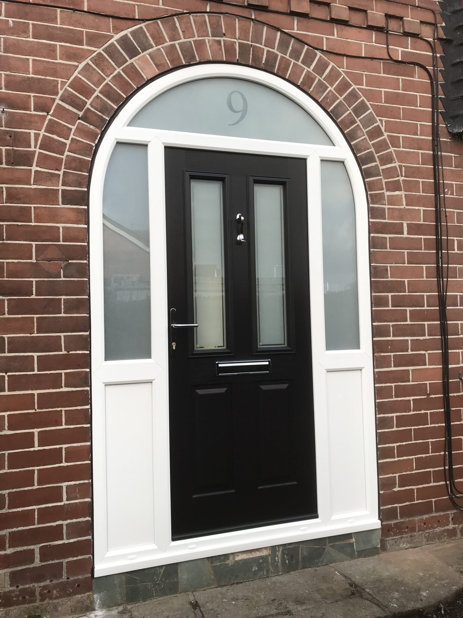 Black composite door with frosted glass panels set in a white frame, beautifully nestled beneath an arched red brick archway. The number 9 is elegantly displayed above the door.