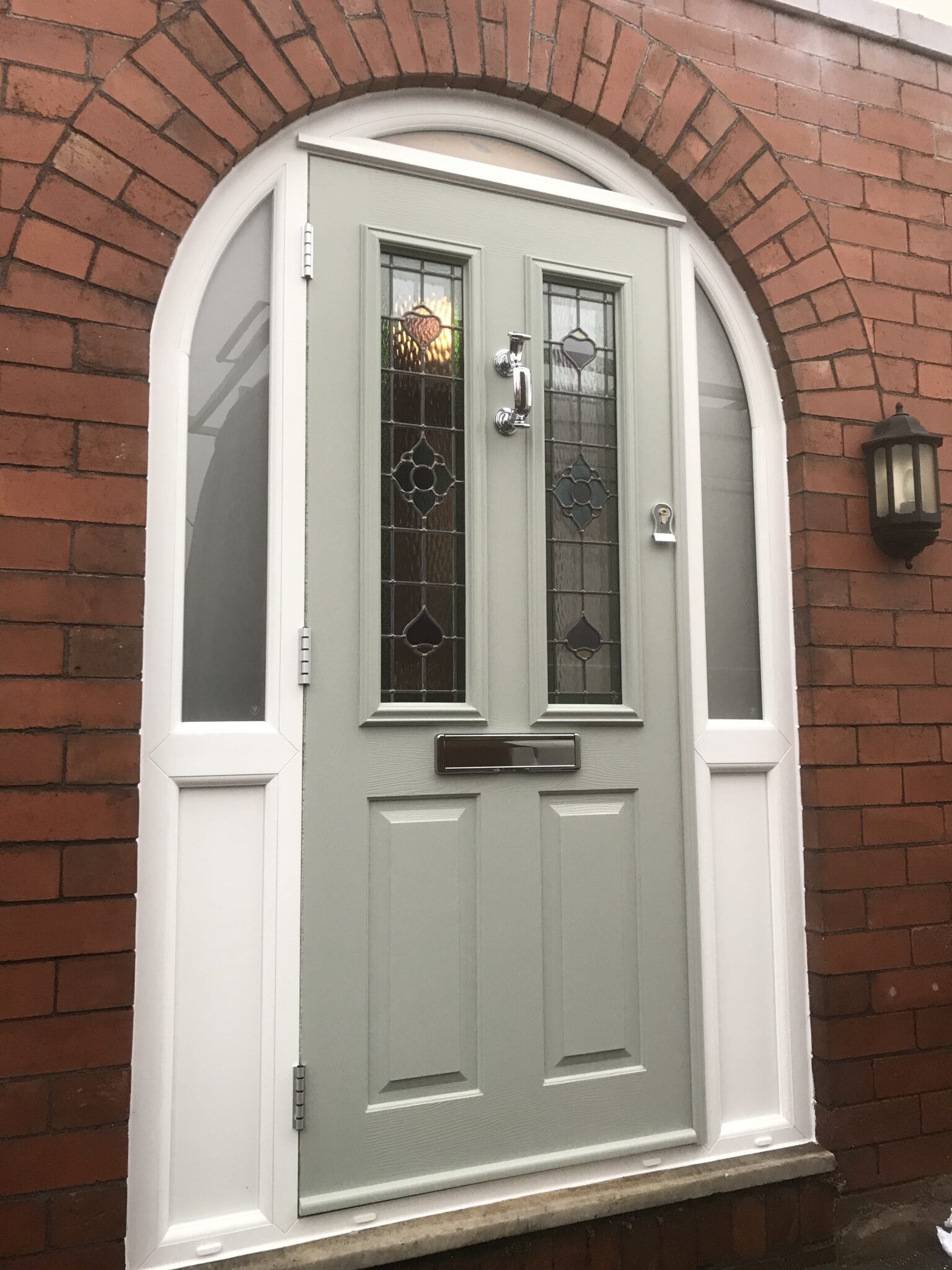 An arched, pale green composite door with decorative glass panels and a chrome handle is set in a red brick archway. To the right, a black outdoor light fixture adds contrast.