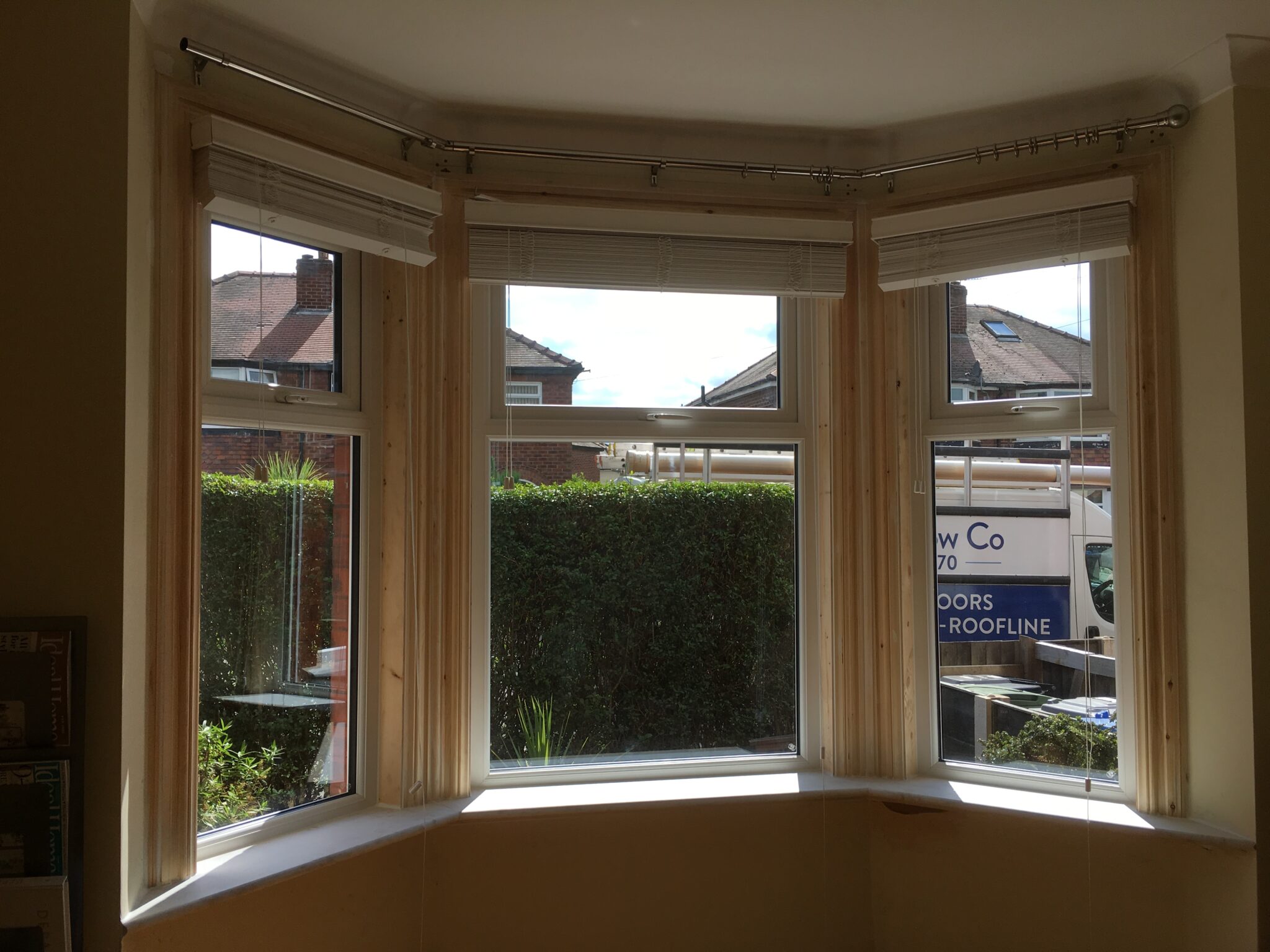 A bay window with four glass panels, featuring flush sash windows and partially open blinds, offers a view of a hedge and parked van outside.