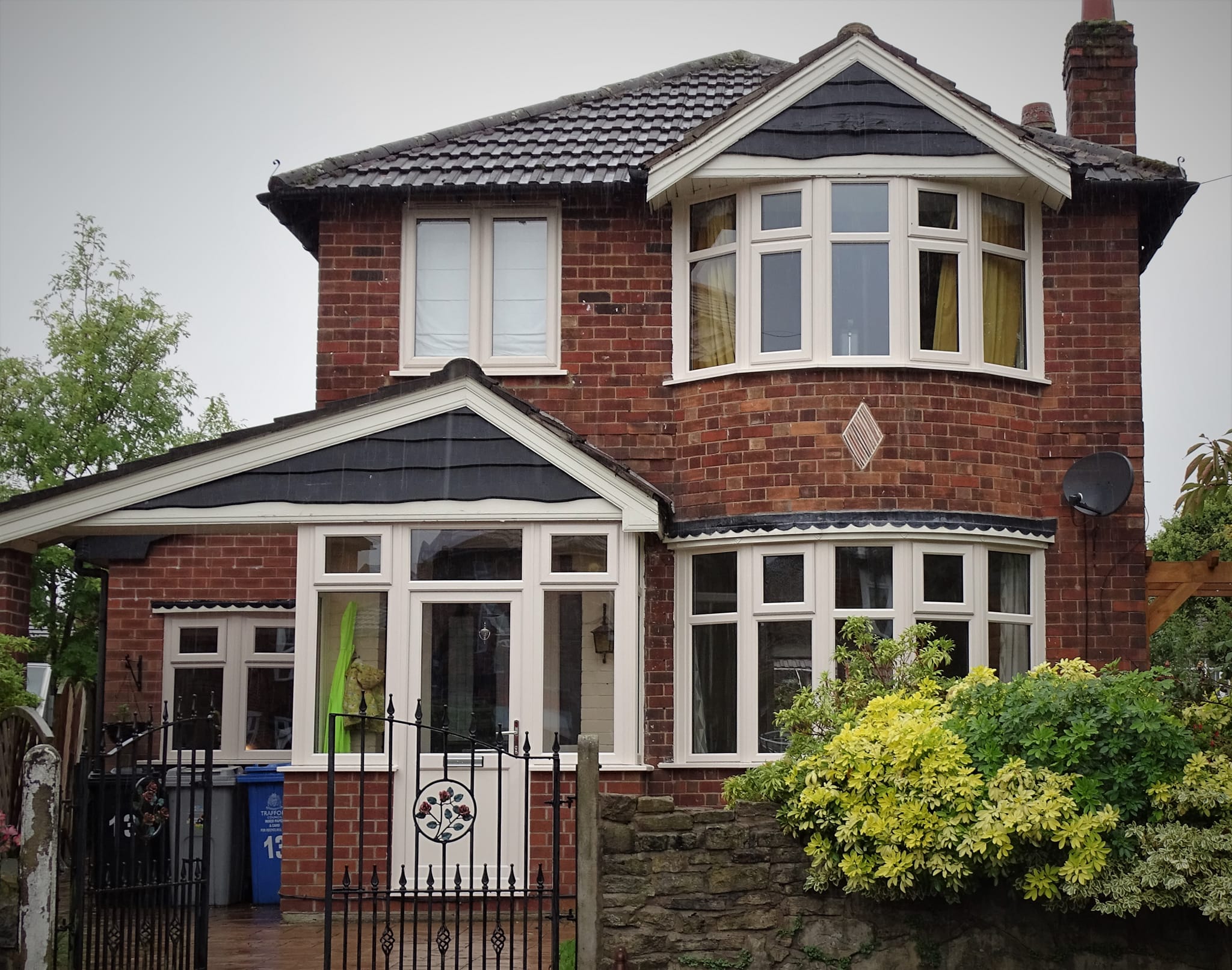 Red brick two-story house with UPVC bay windows, a small front gate, and shrubs in the garden. A satellite dish is attached to the side wall.