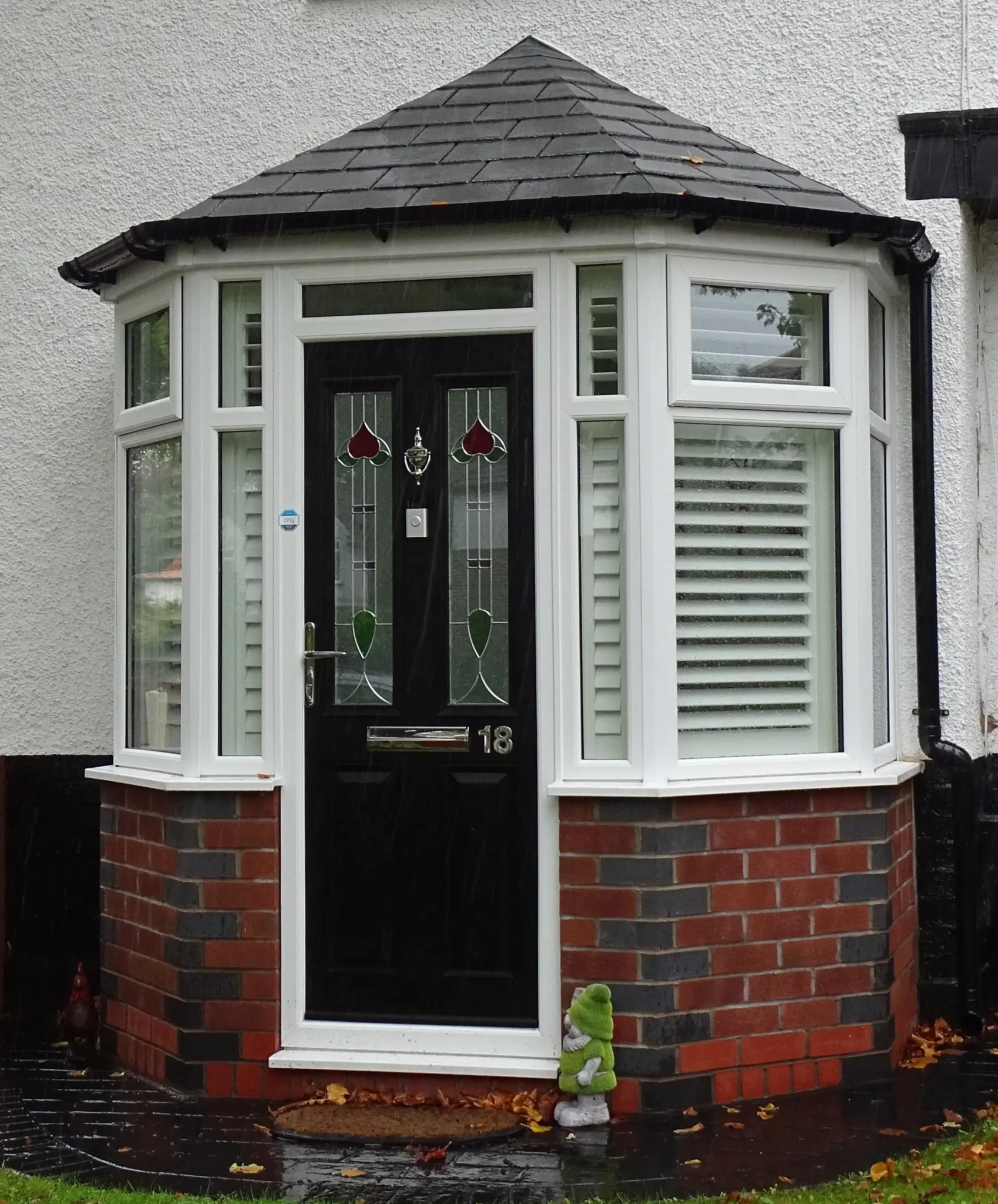 Bay-front door with black frame, stained glass panels, and house number 18. Red brick base, white walls, and a small frog statue near the entrance.