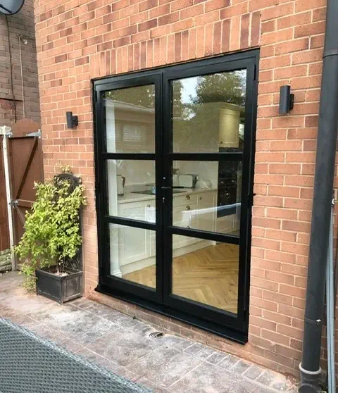 A brick wall with a large black-framed window, reminiscent of French doors, reveals a white kitchen interior. Two wall lights flank the window, and a potted plant sits to the left.