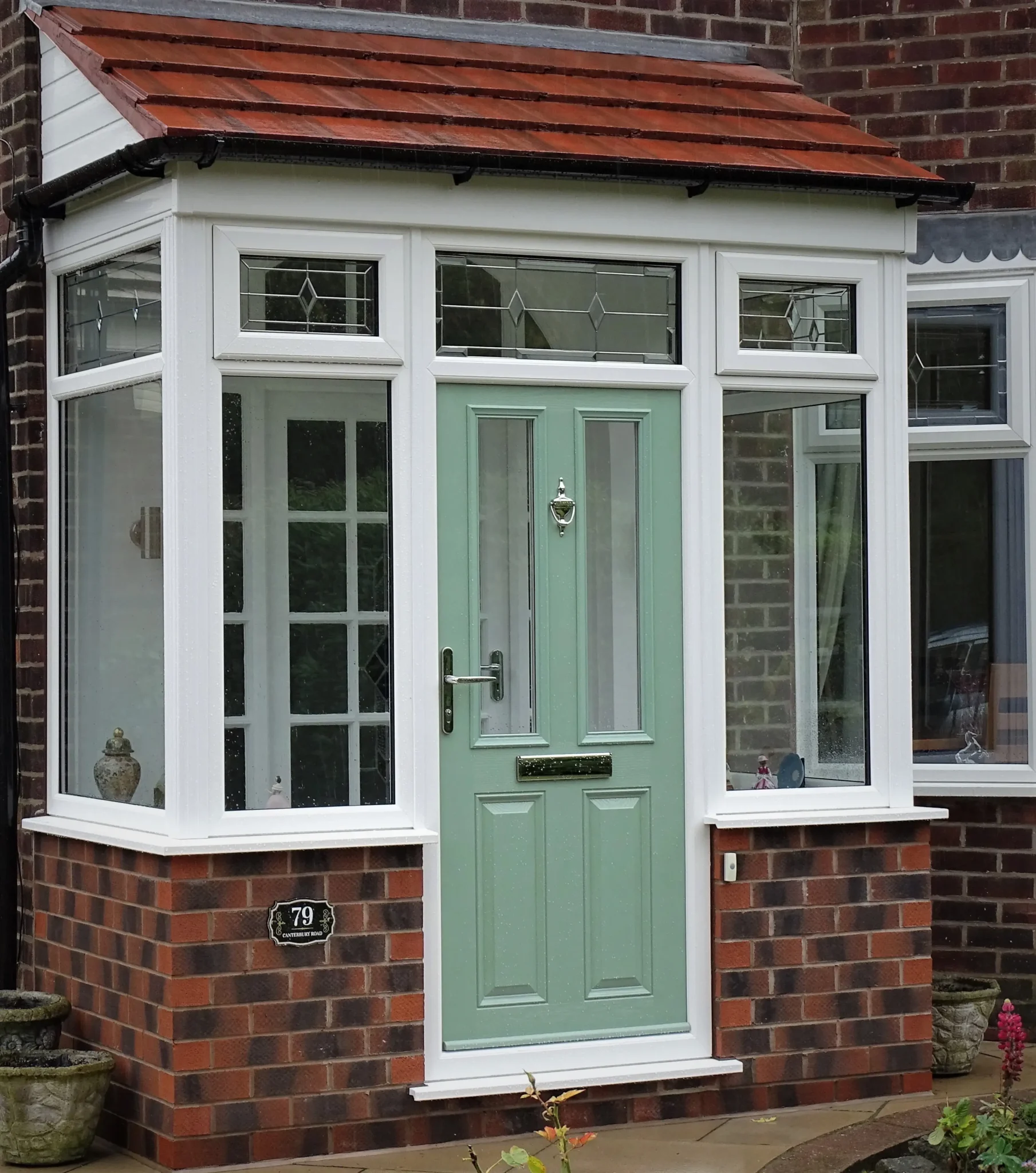 A house entrance with a light green door, flanked by large windows and a small porch with a red-tiled roof.