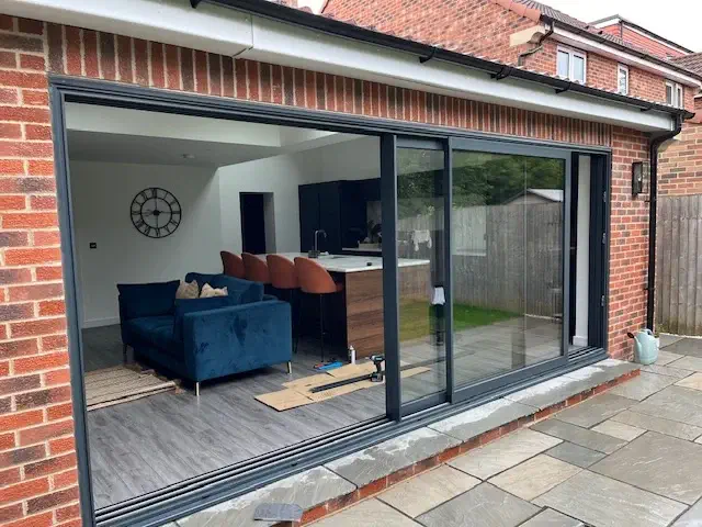 Modern interior featuring a blue sofa, bar stools, and a kitchen visible through large sliding patio doors of a brick house. Patio in the foreground.